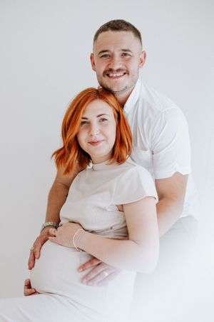 A couple dressed in white embracing for a maternity photo against a light background.