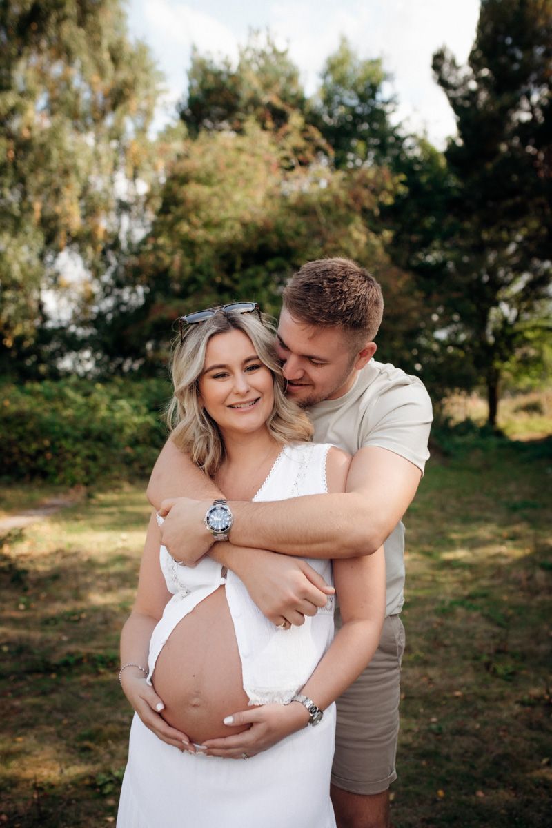 Intimate maternity photo session showing couple embracing in natural outdoor setting with trees in background.