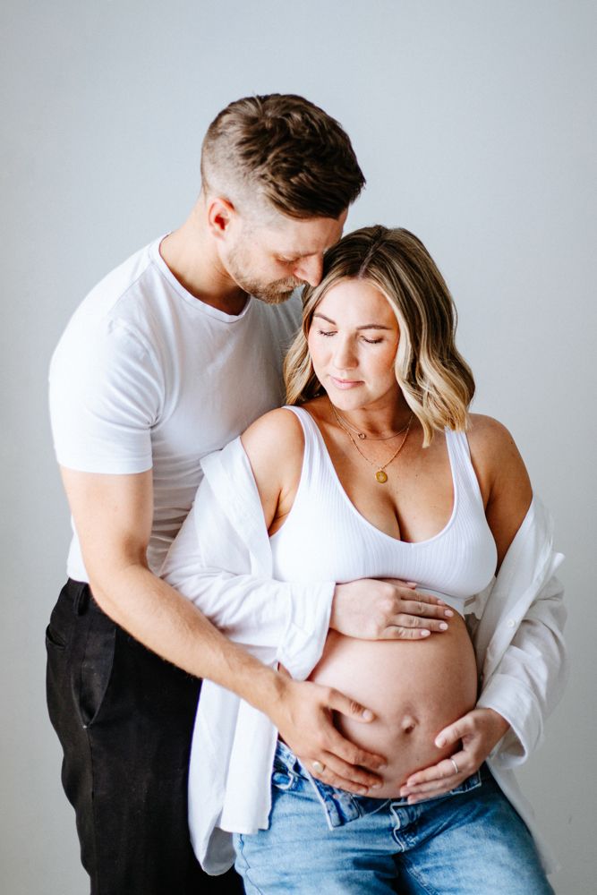 Pregnant couple in casual white clothing share an intimate moment against light grey background during maternity photoshoot.