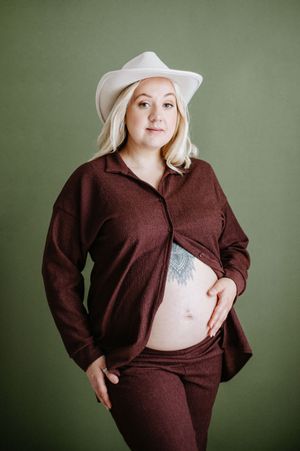 Portrait of someone in a white cowboy hat and brown outfit showing a belly tattoo against a green background.