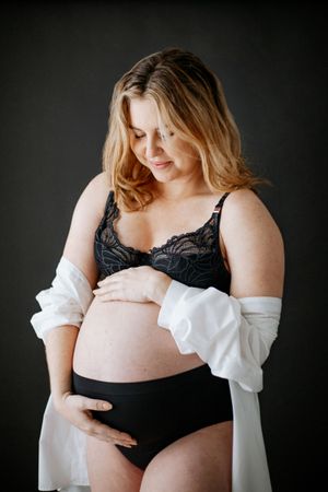 Intimate during maternity photoshoot in Nottingham with pregnant woman wearing black lingerie and white cardigan against dark background.
