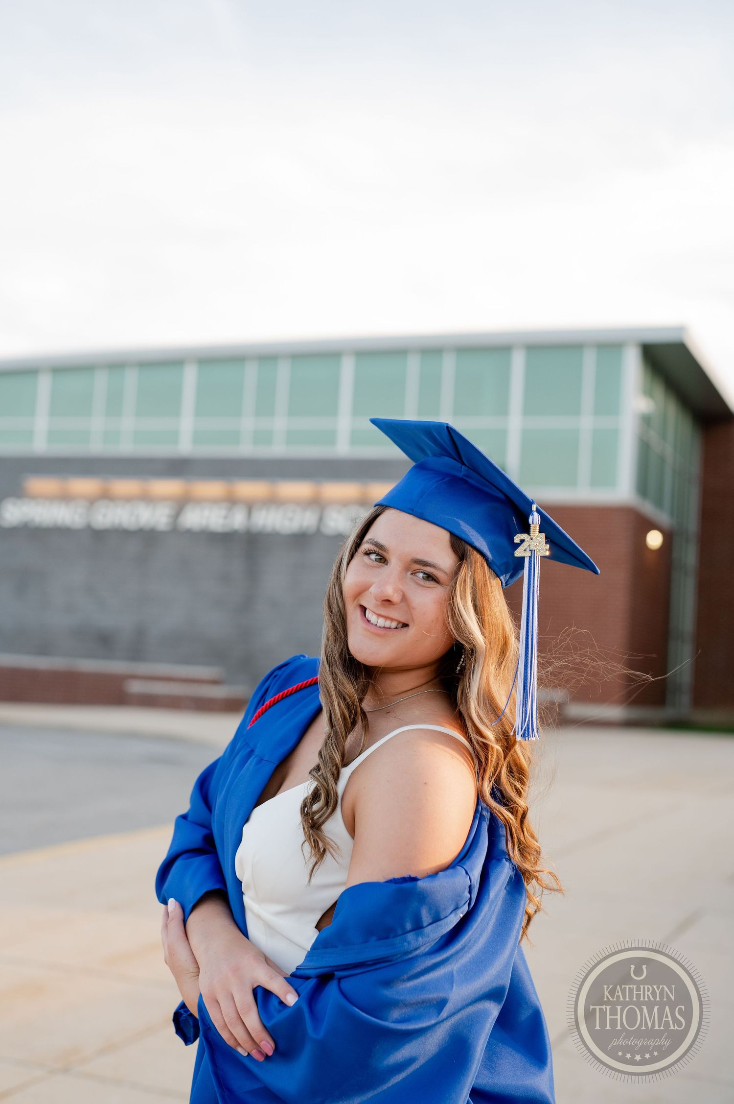 Graduate in blue cap and gown smiles while posing against school building exterior.