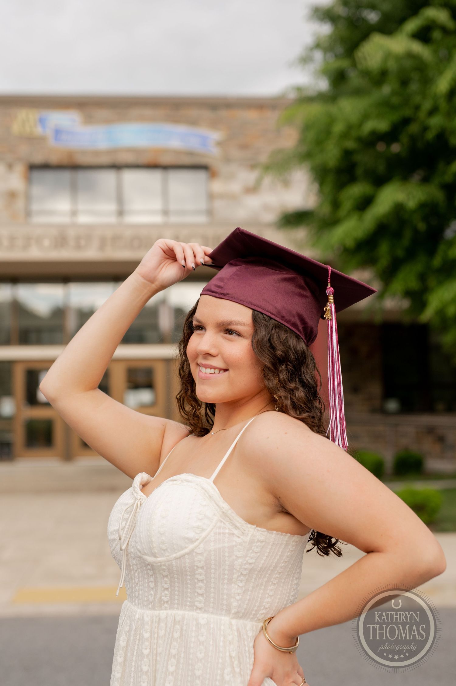 Graduate adjusts maroon cap while wearing white dress outside school entrance.