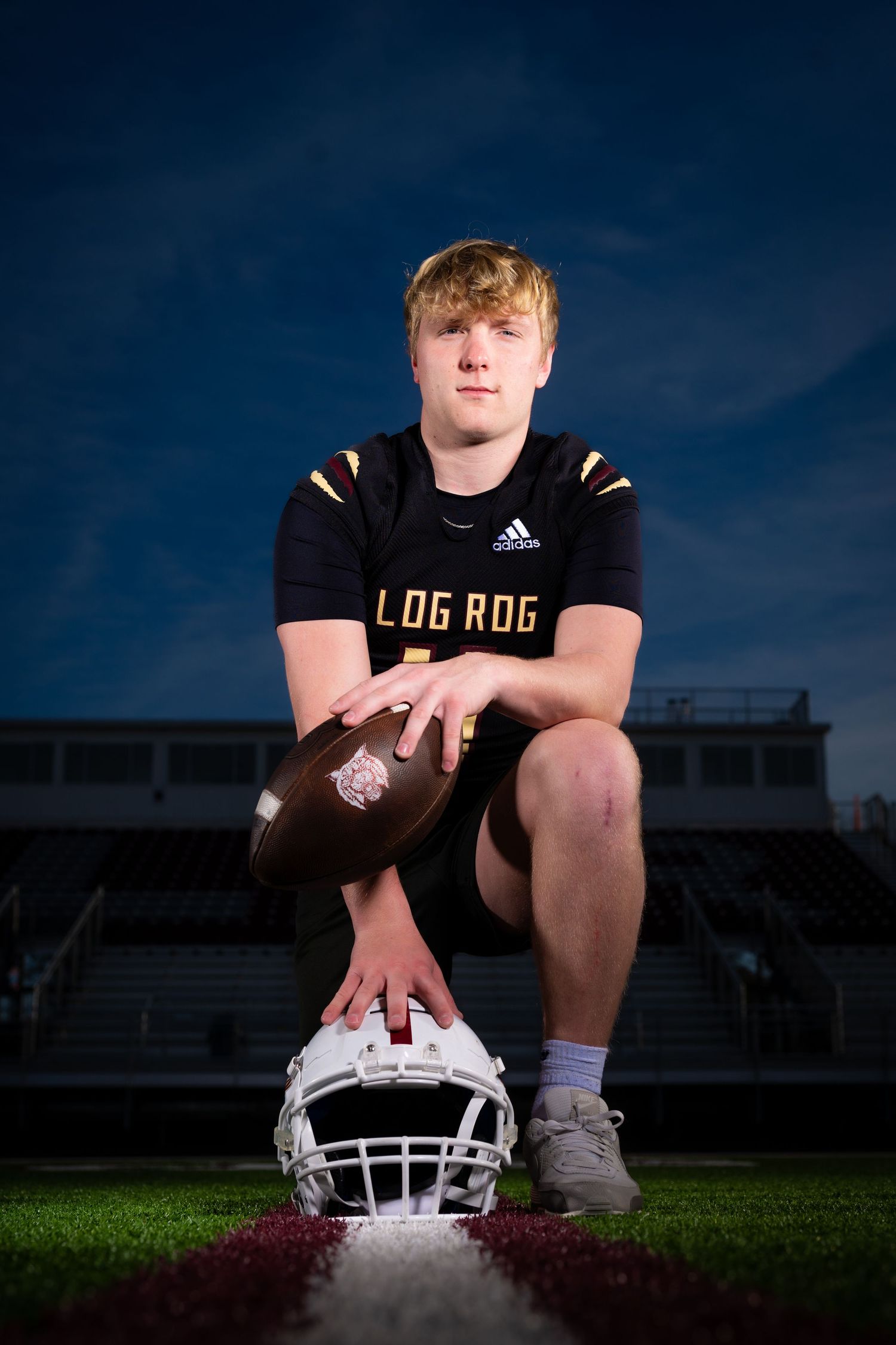 Football player kneeling with ball and helmet on field at dusk with stadium lights.