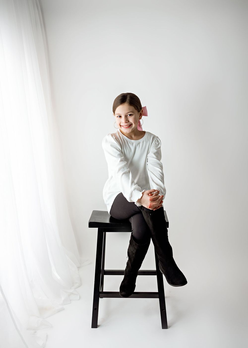Child in white long sleeve shirt sits playfully on black wooden stool against white studio backdrop.