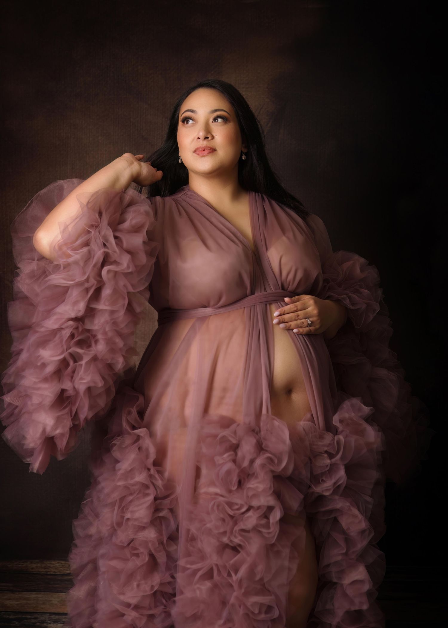 Dramatic maternity portrait in dusty rose ruffled gown against dark background with soft lighting and ethereal styling.