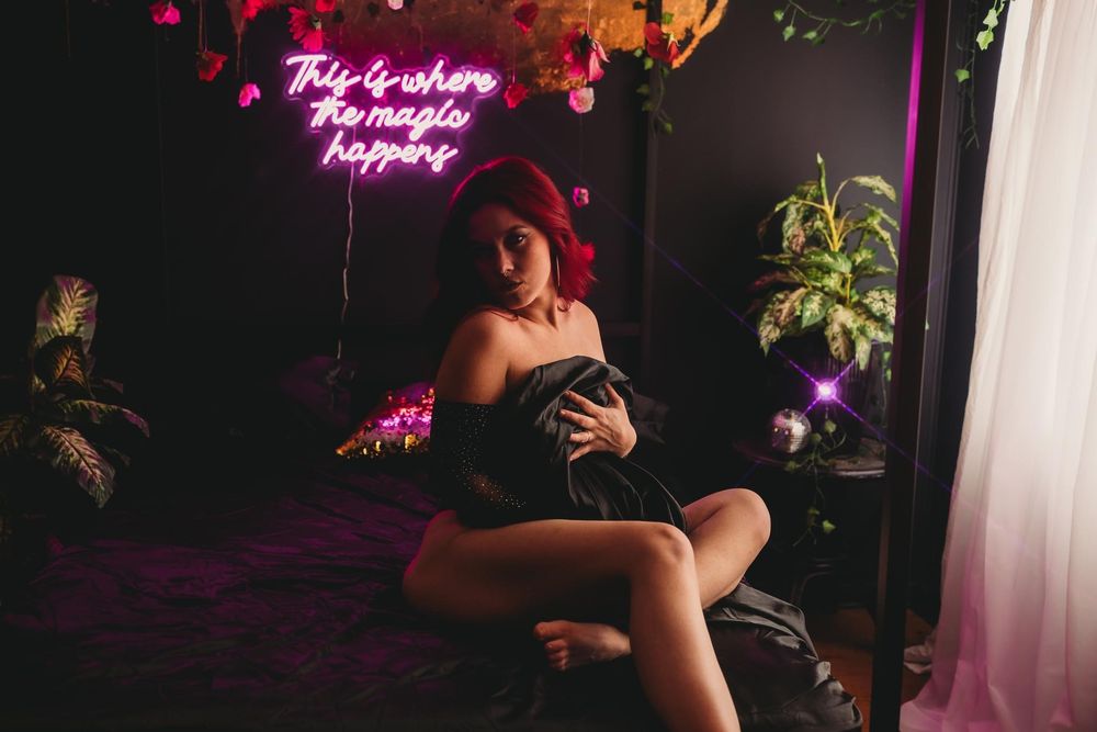 Silhouetted figures dance and pose in a dark room illuminated by pink neon lights and lush greenery.
