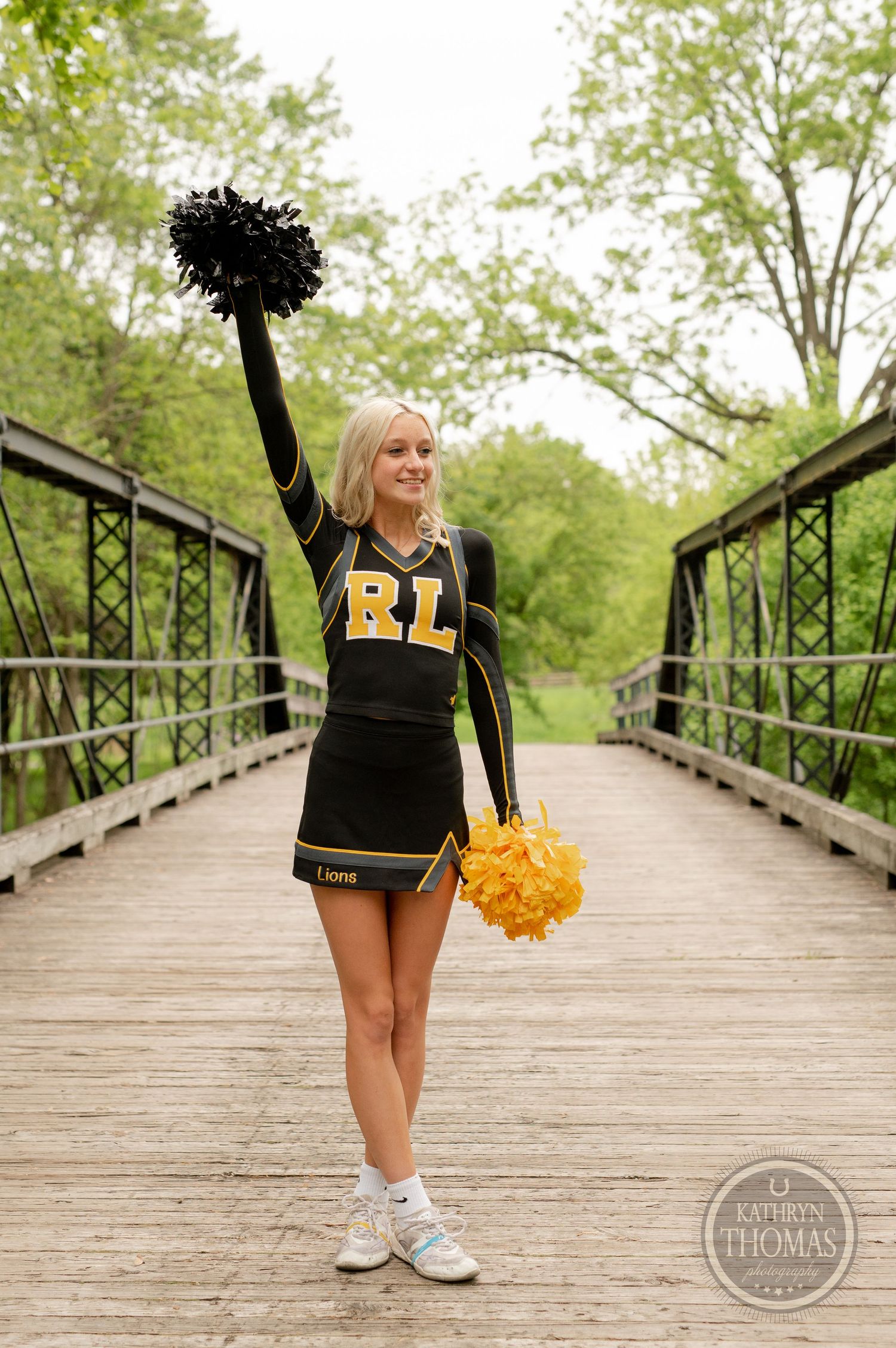Cheerleader in black uniform standing with pom poms on wooden bridge.