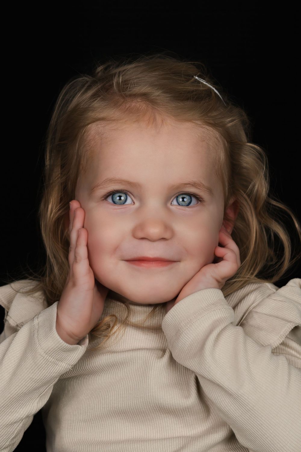 Een klein meisje dat lachend in de camera kijkt. Ze heeft haar hand onder haar kin en haar andere hand tegen haar wang. Lichte kleding.