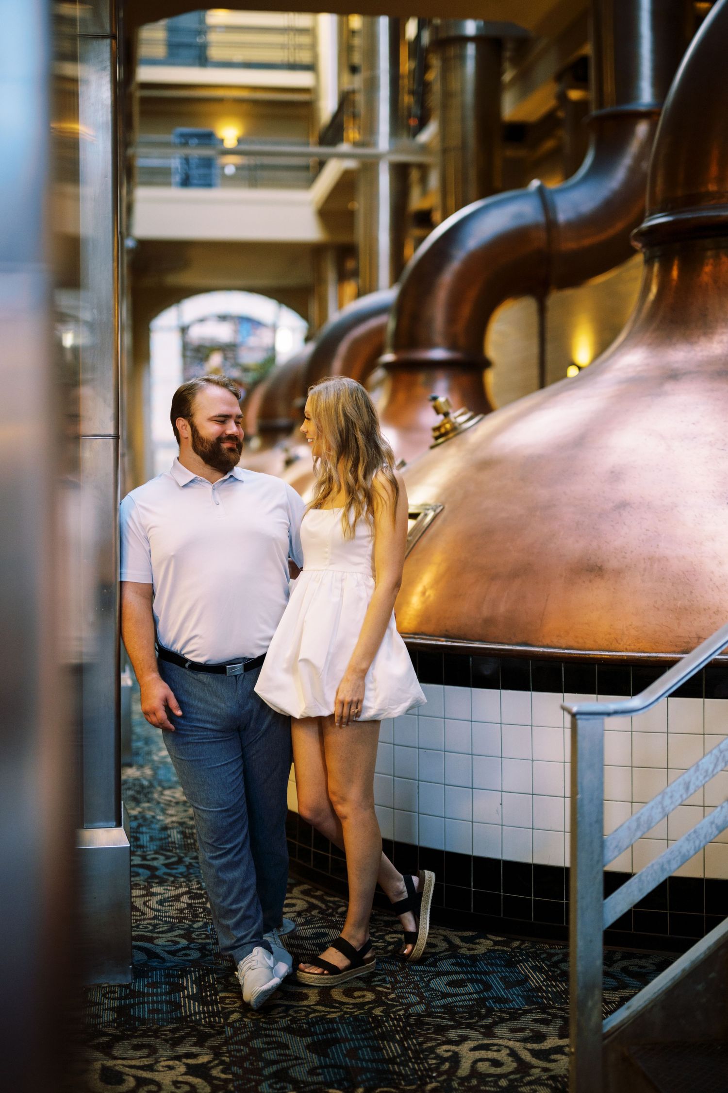A couple tours a brewery or distillery facility with large copper stills and industrial equipment in the background.