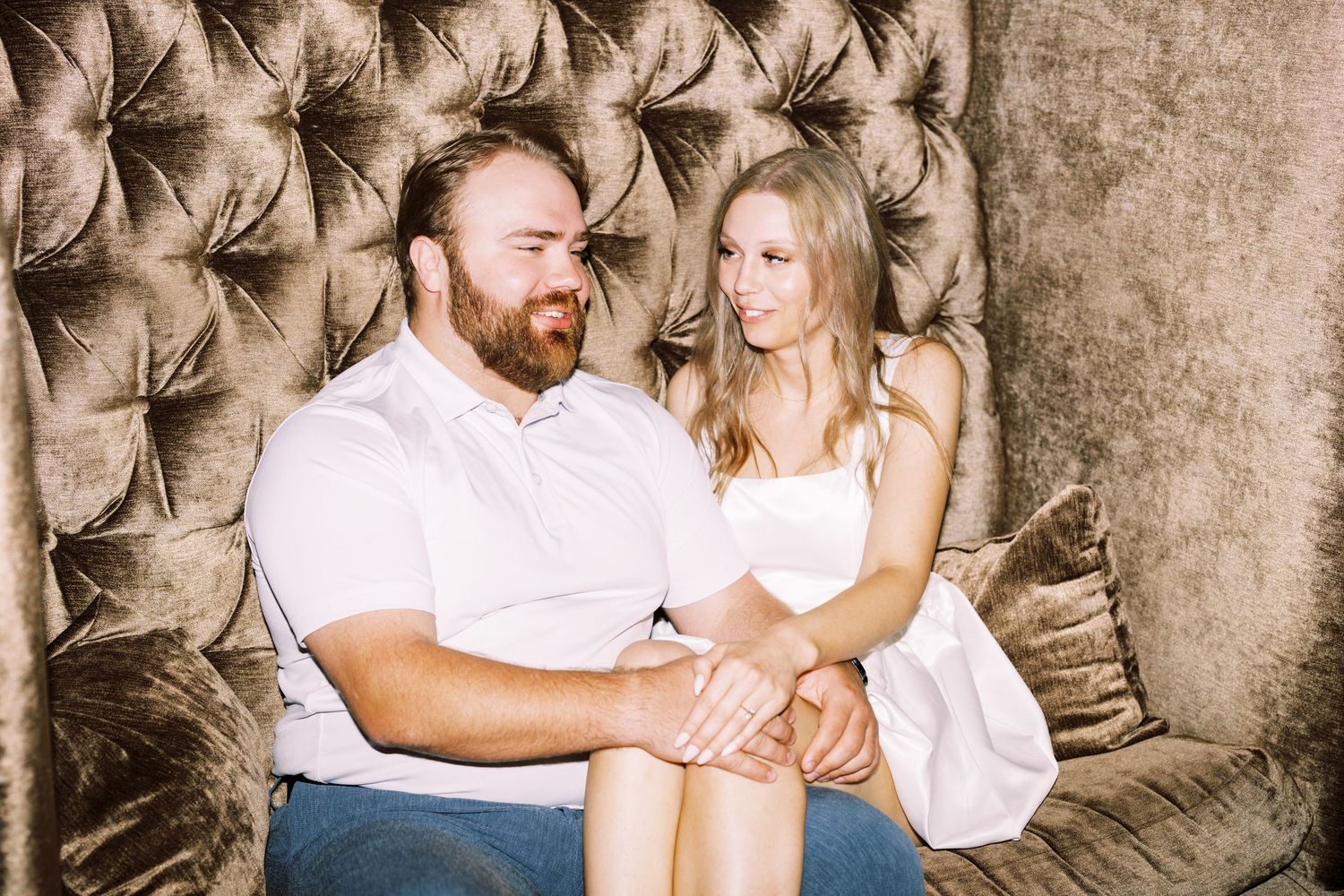 A couple sits together intimately on a brown tufted velvet couch in a romantic portrait setting.