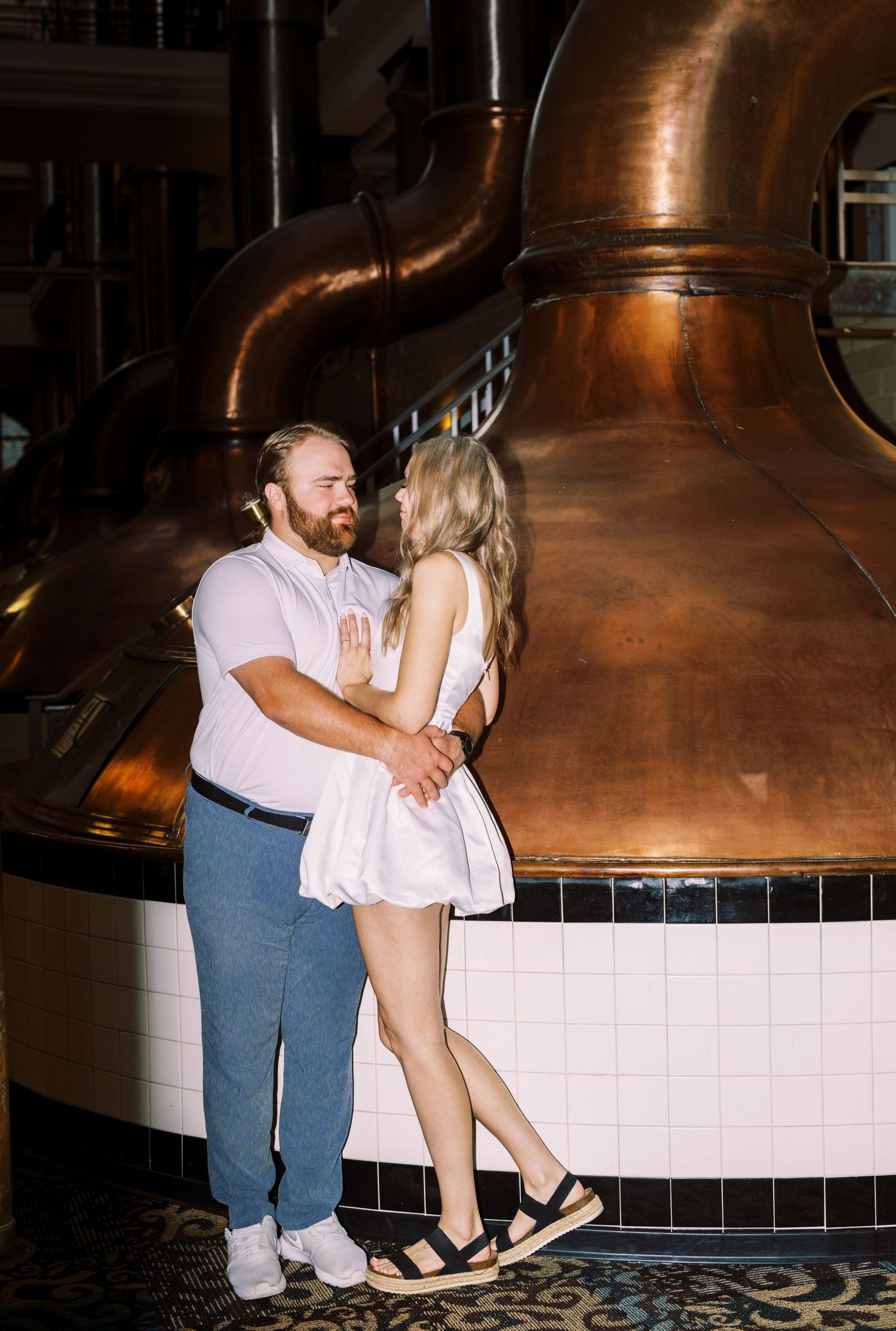 A romantic couple embraces in front of a large copper brewing tank in a brewery.