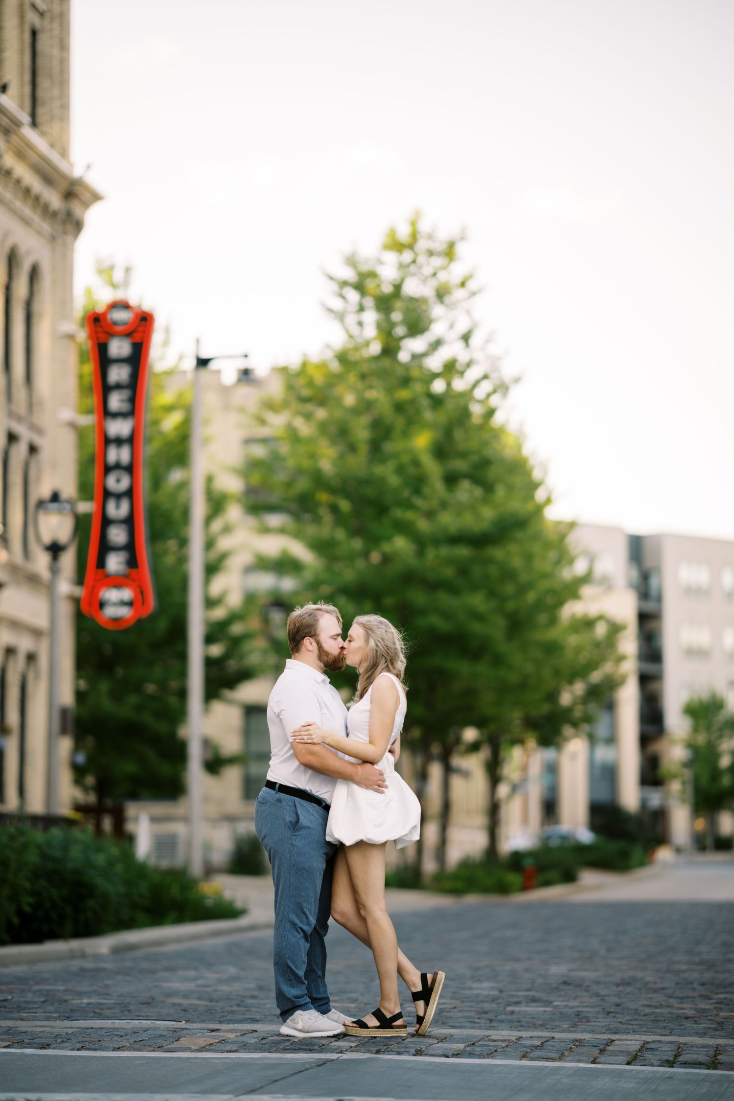 A romantic couple shares a tender moment in front of a red neon sign on an urban street corner.