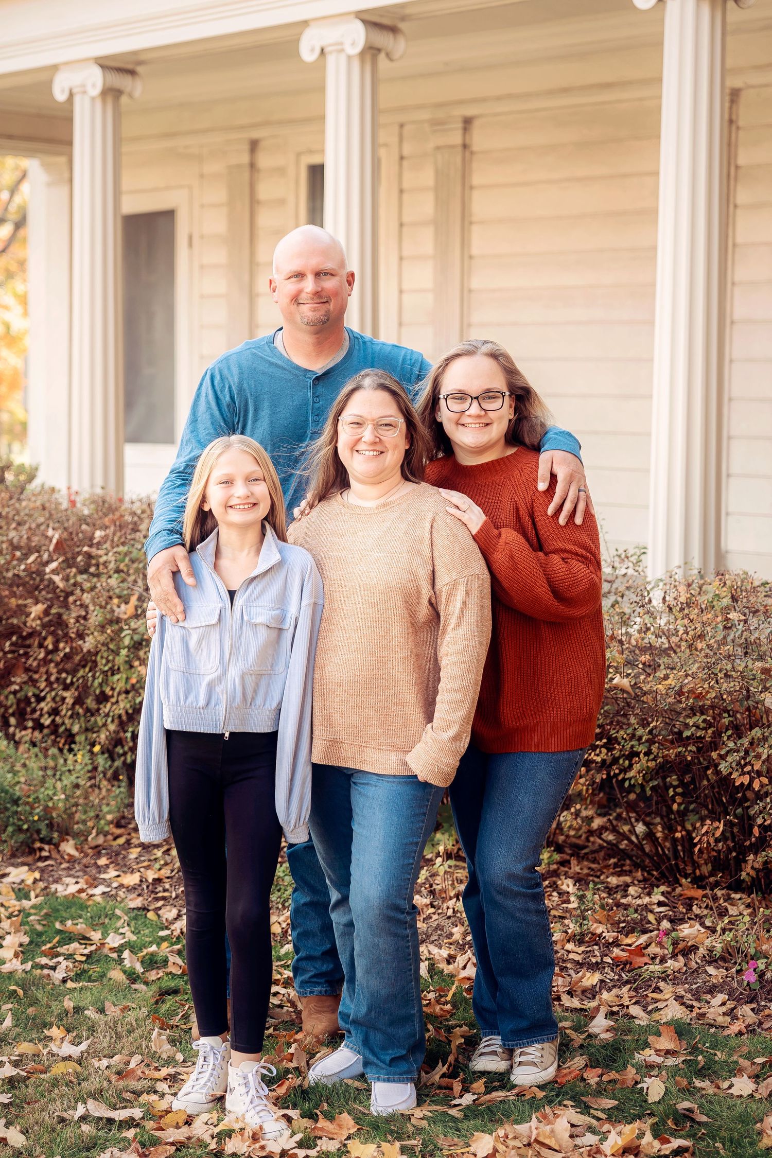 Family of four poses in front of white pillared porch during autumn with fallen leaves on ground-Michigan Detroit Novi Livonia Photographer