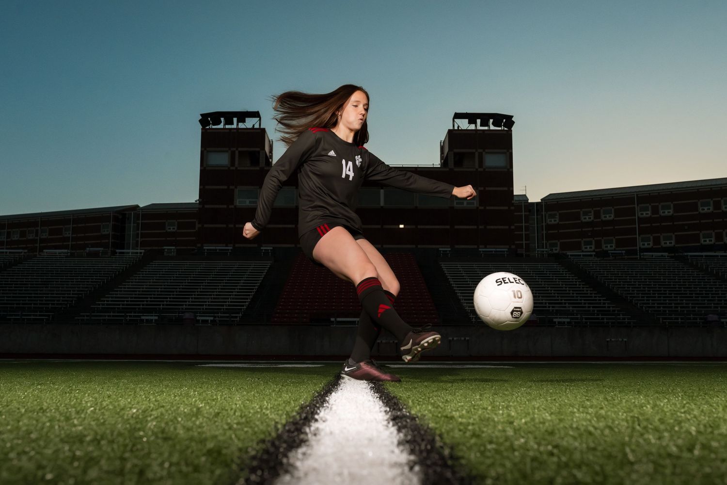 Soccer player kicks ball on field with stadium seating in background at dusk.