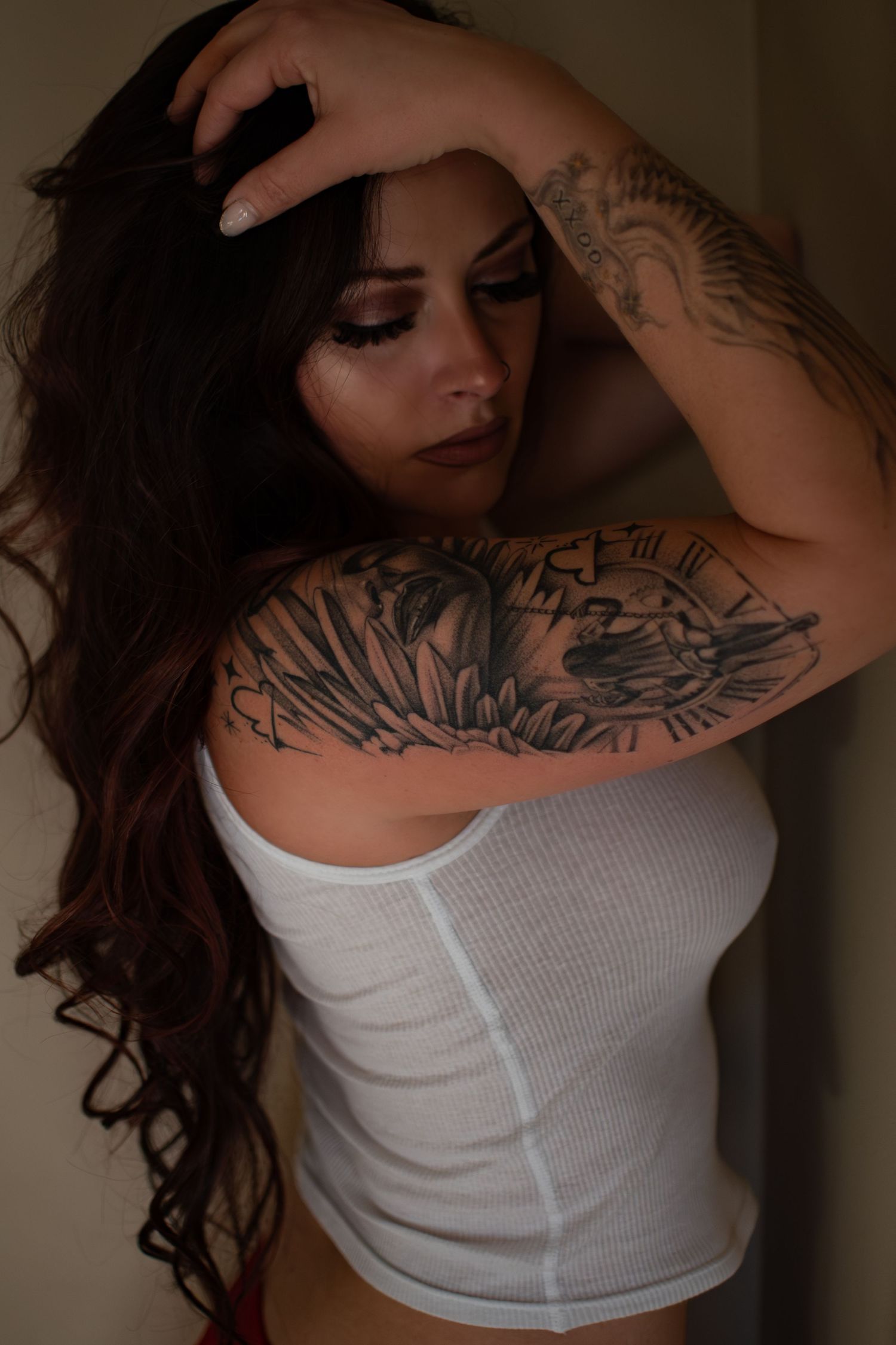 Woman with long dark hair and artistic tattoos poses in white tank top against dark background.