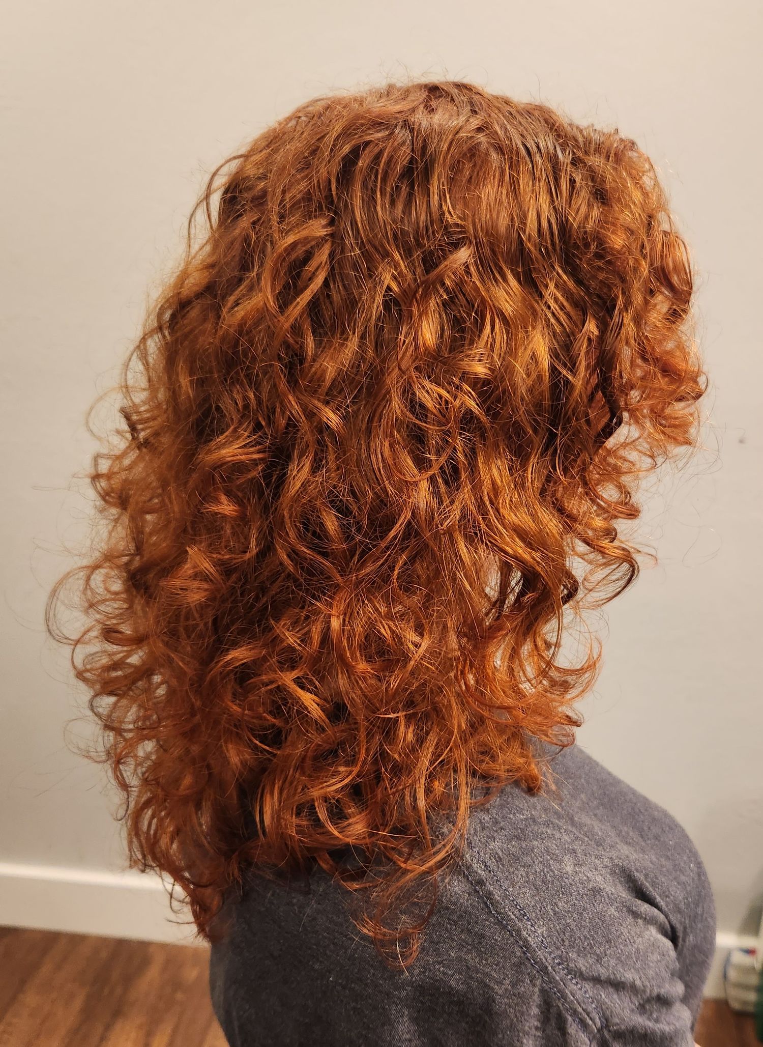Back view of a woman with voluminous curly auburn hair against a gray background.