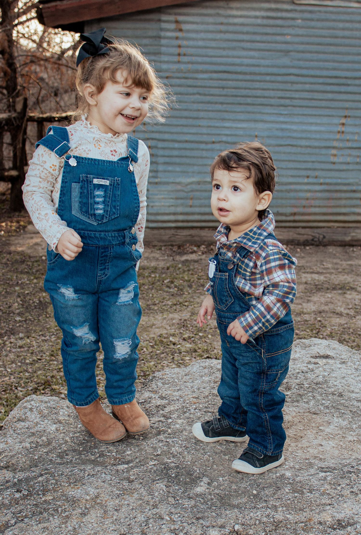 Two children wearing matching denim overalls and plaid shirts stand together outdoors by a metal wall.