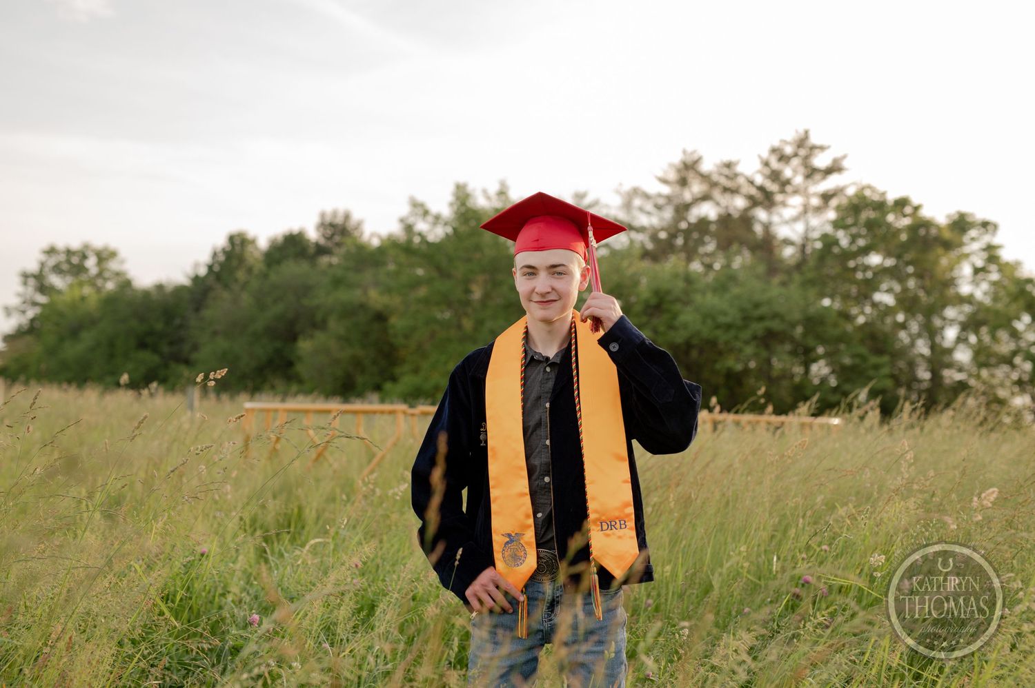 Graduate in red cap and gold stole smiles in grassy field during sunset photo session.