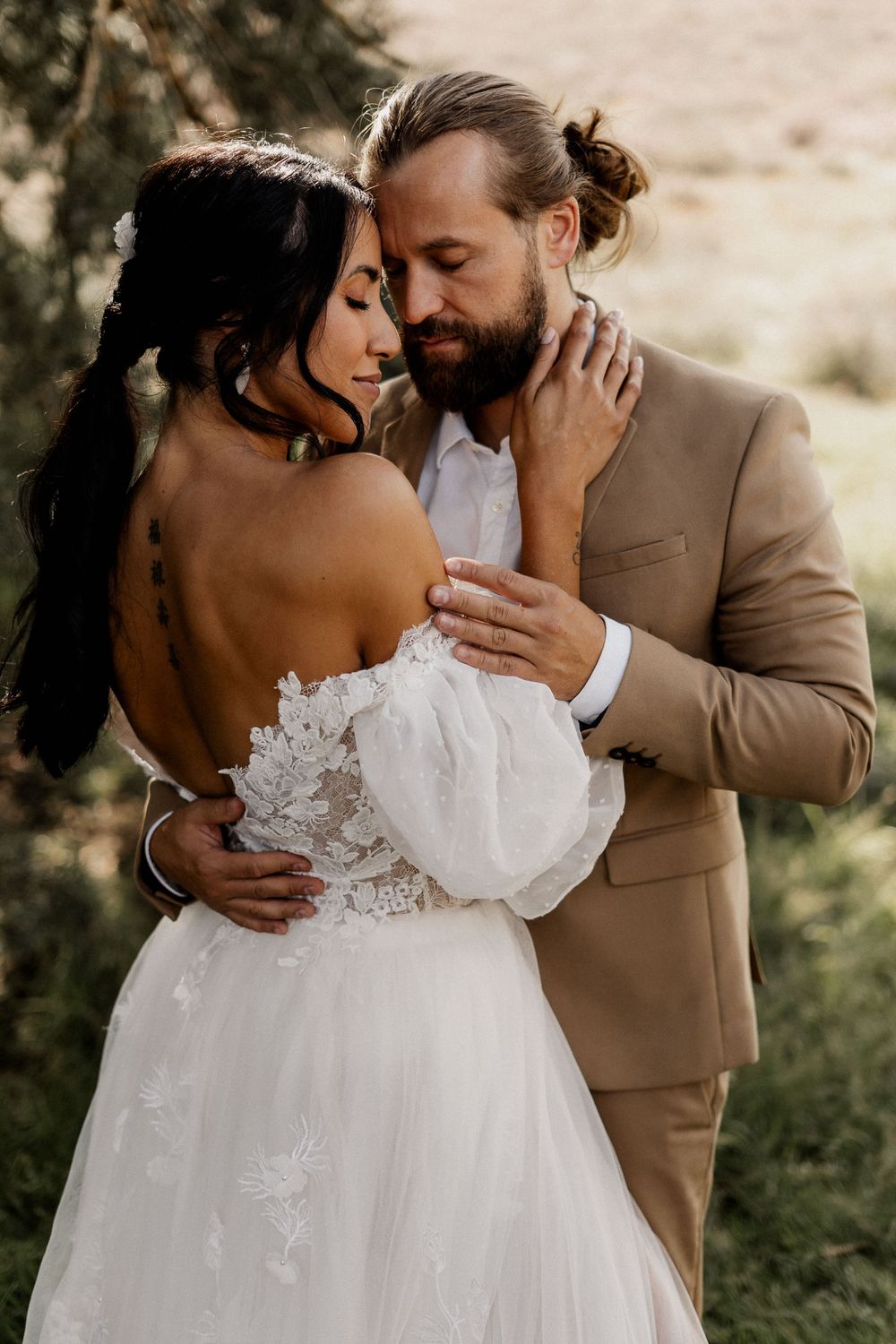 Intimate wedding photo of a couple embracing in warm sunlight, bride in white lace gown and groom in tan suit.