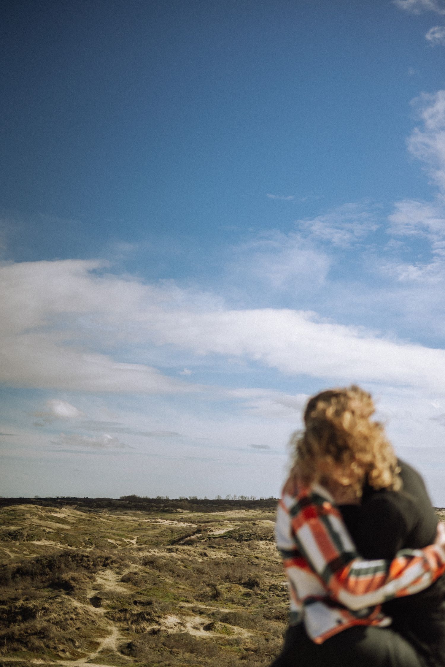 A figure in a striped sweater sits overlooking a vast coastal landscape with cloudy blue skies.