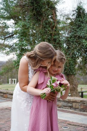 A tender moment captured outdoors as two people in white and pink dresses share flowers in a garden setting.