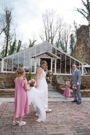 Wedding party gathered on brick patio in front of elegant greenhouse venue with stone walls and rustic chimney.