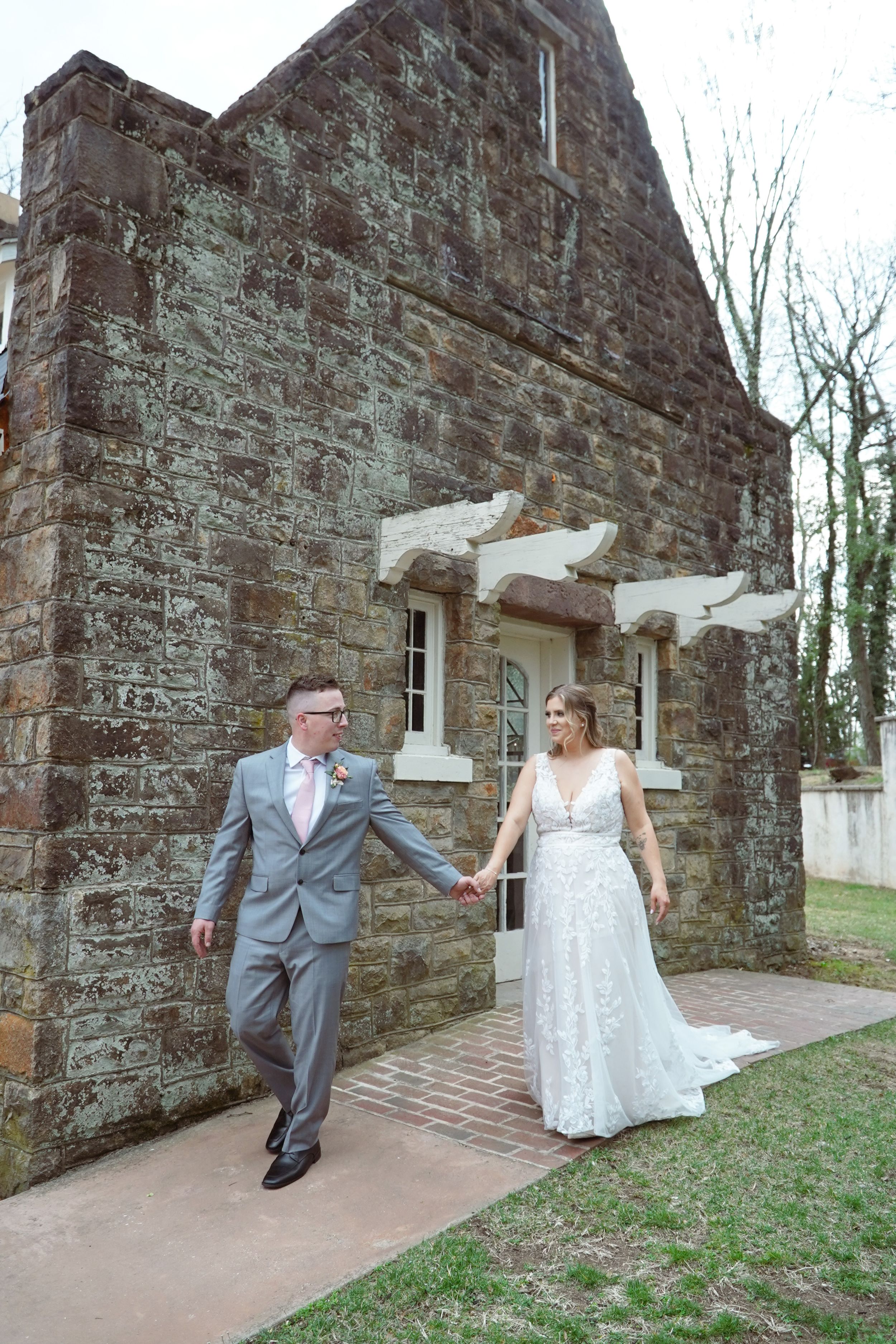 A couple in formal attire walks hand-in-hand along a stone pathway next to a historic building with rustic walls and windows.