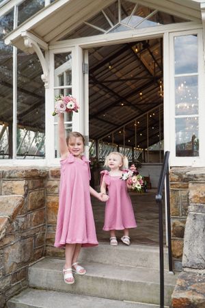 Two young children in matching pink dresses stand on stone steps at the entrance of a rustic building with string lights.