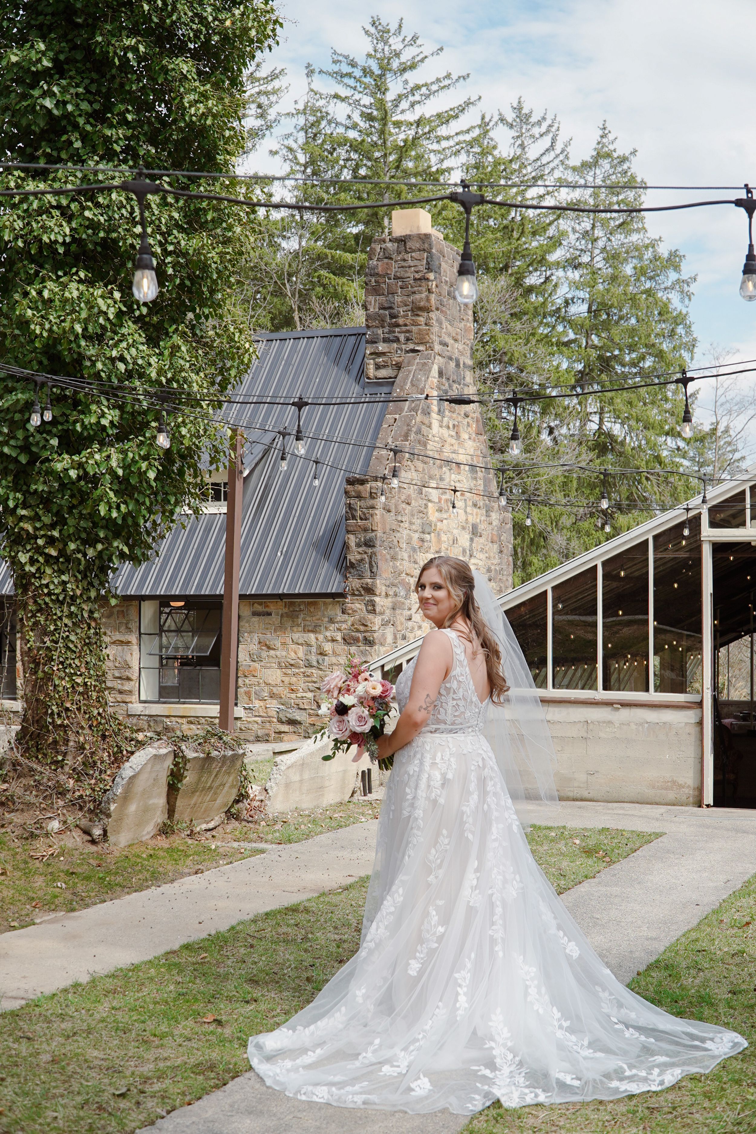 Beautiful wedding dress with lace details and long train photographed outside rustic stone building with metal roof.