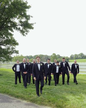 Groomsmen in black tuxedos walking on grass path with white fence in background.