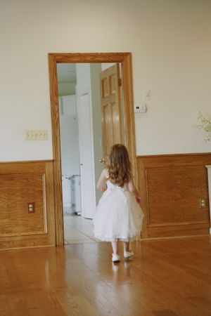 Young flower girl in white dress walks toward wooden doorway in sunlit hallway.