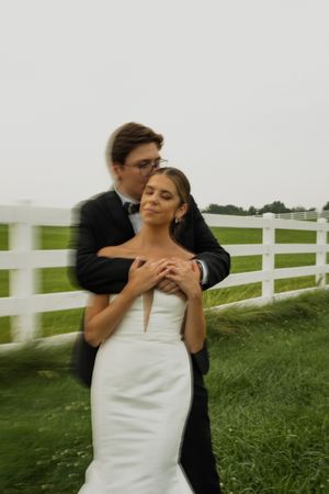 Romantic wedding portrait by white fence with couple embracing in formal attire.