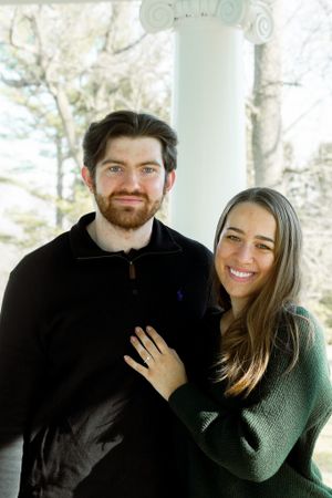 A newly engaged couple poses for a portrait under a white column showing off the ring.