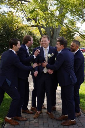 Groomsmen in navy suits playfully posing with groom on brick pathway under trees.