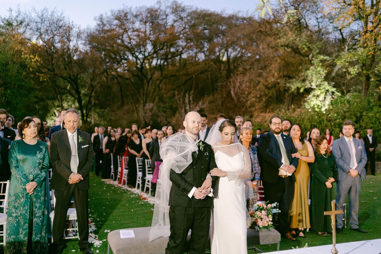 An outdoor wedding ceremony with guests standing in rows among trees at dusk.