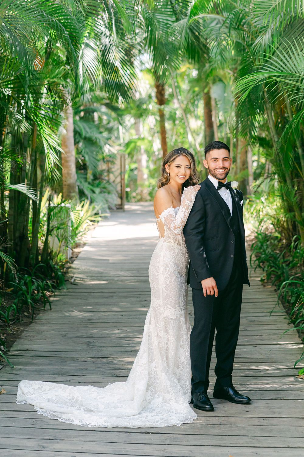 A wedding couple poses on a wooden walkway surrounded by lush tropical palm trees.