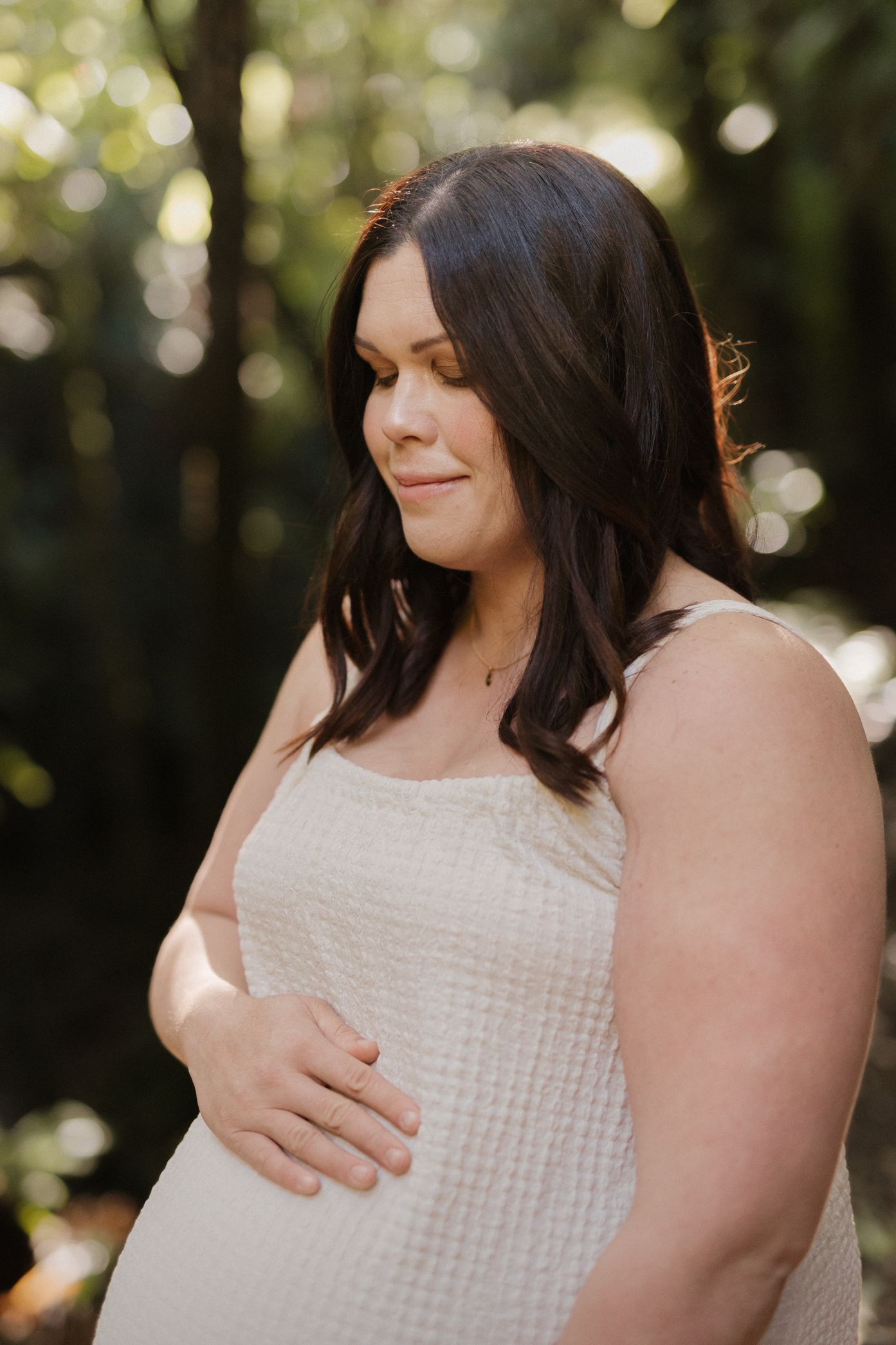A woman in a white textured dress holds her baby bump against a sunlit forest backdrop.