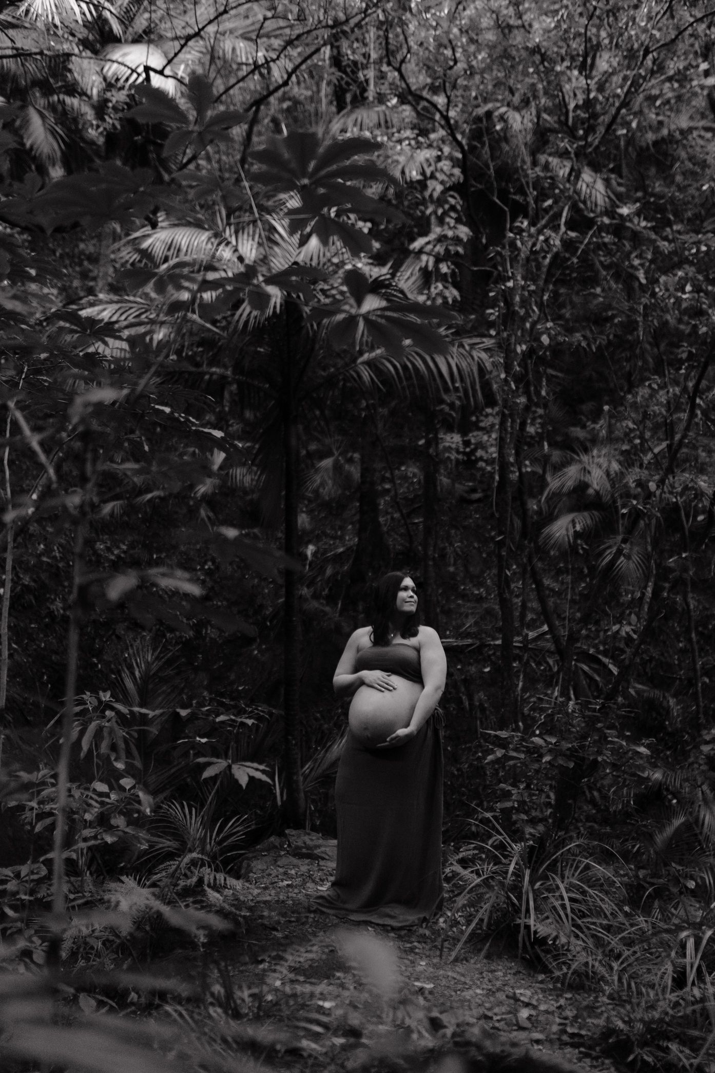 A black and white photograph of an expectant mother standing in a lush tropical forest setting.
