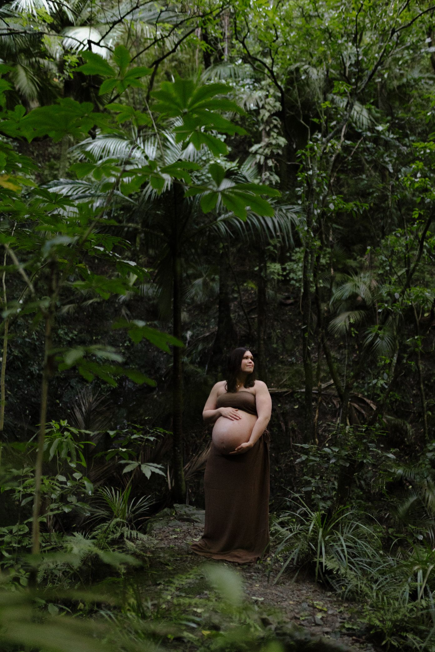A person in a brown dress sits contemplatively among lush tropical foliage in a forest setting.