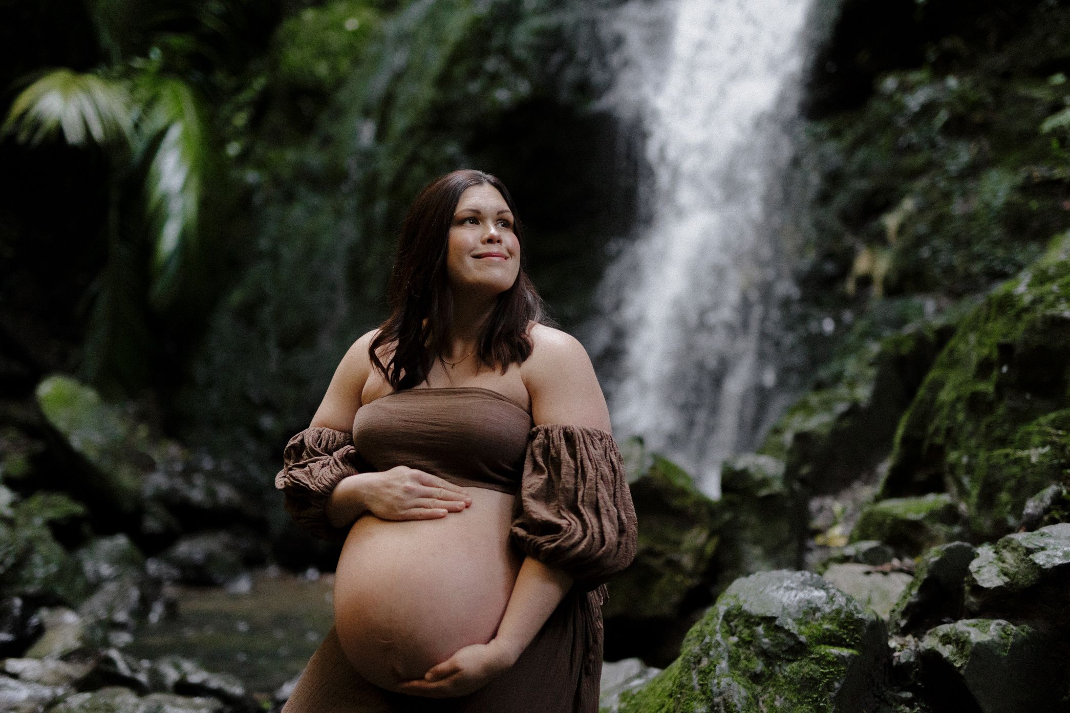 A maternity photoshoot captures an intimate moment against a scenic waterfall backdrop in a lush forest setting.
