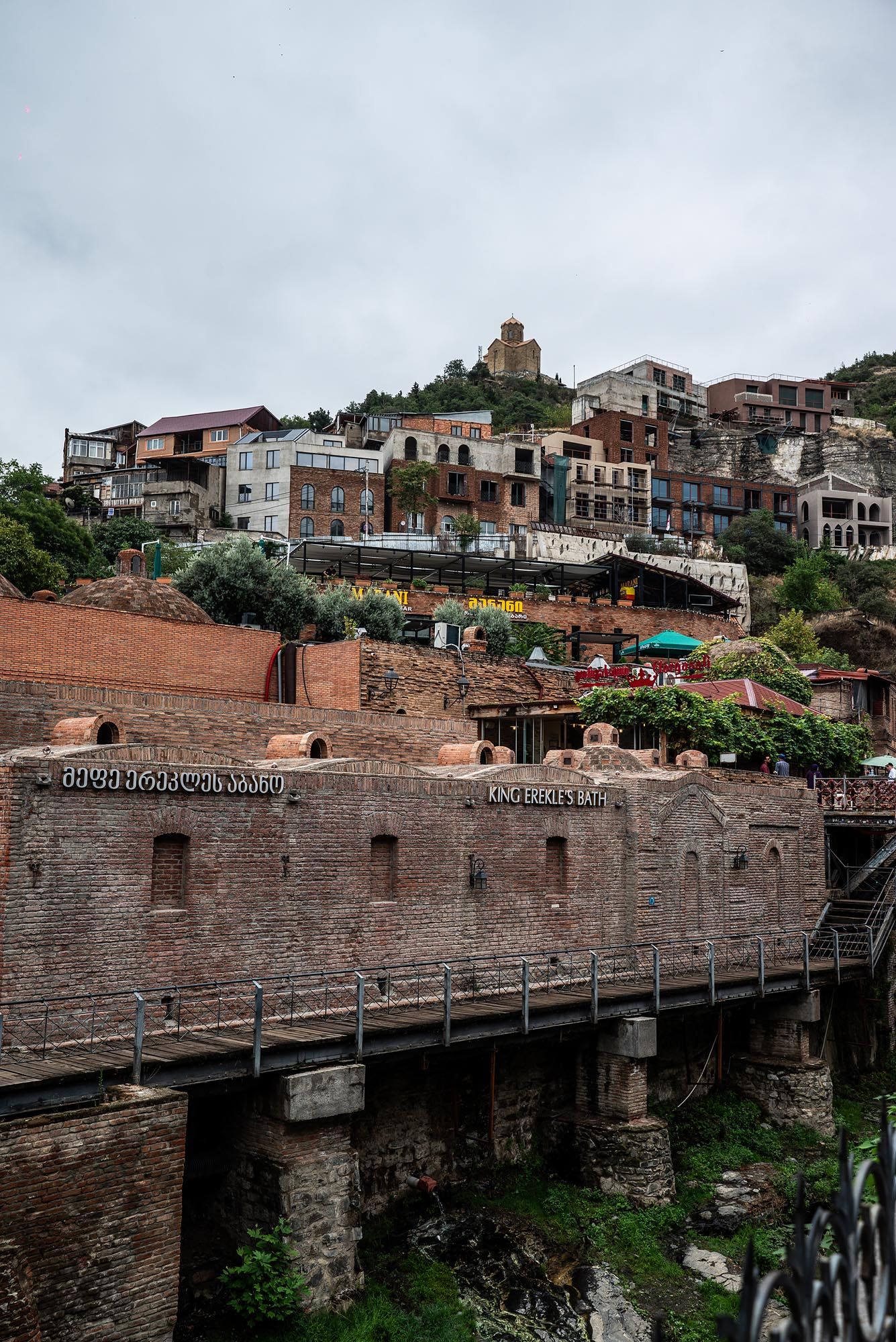 Historic stone wall and bridge structure with residential buildings on hillside in Tbilisi, Georgia.