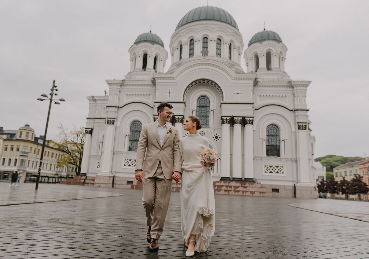 Couple in wedding attire standing before a grand white Orthodox church with domed architecture.