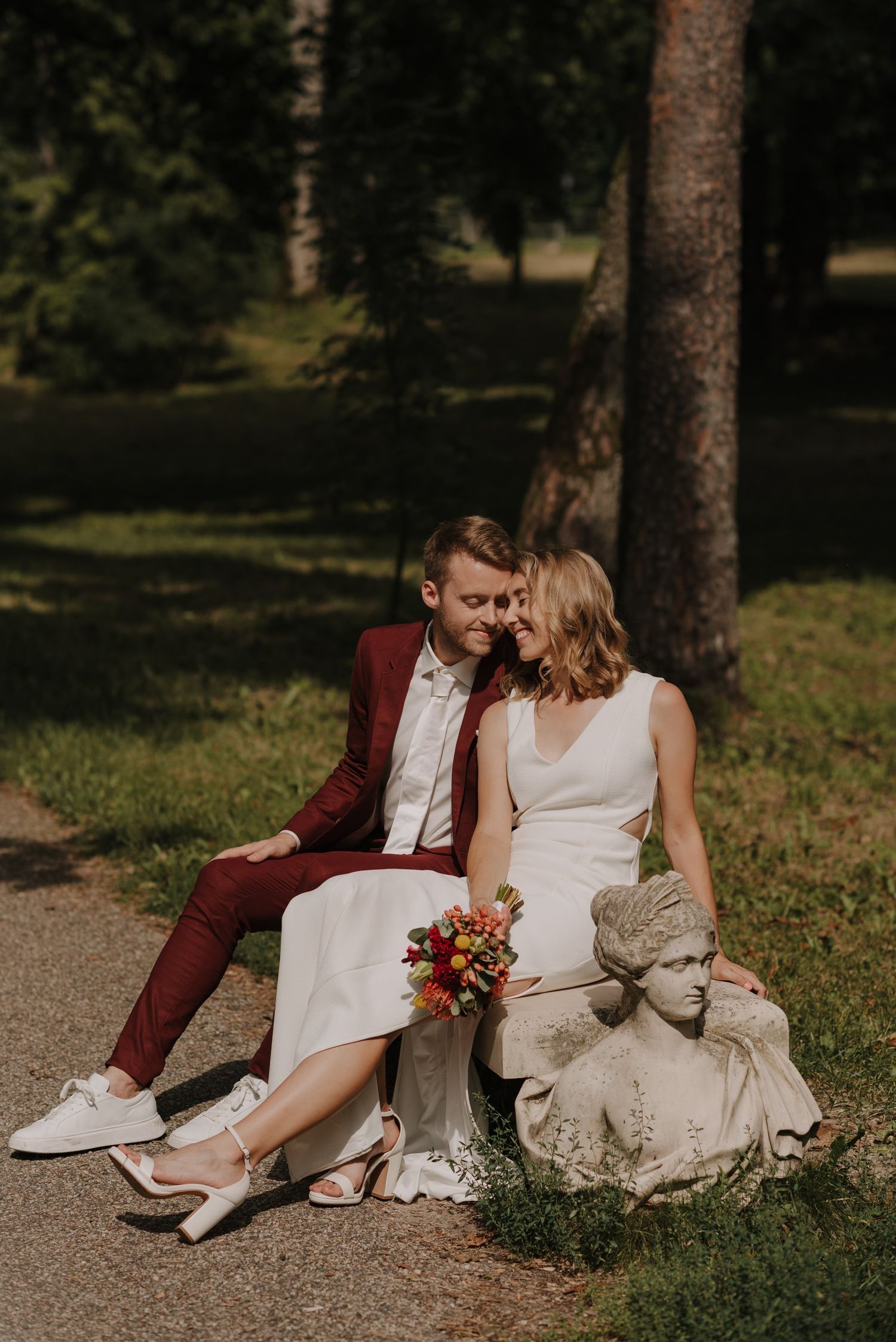 A couple sits together on a garden path during their wedding photoshoot wearing burgundy and white.