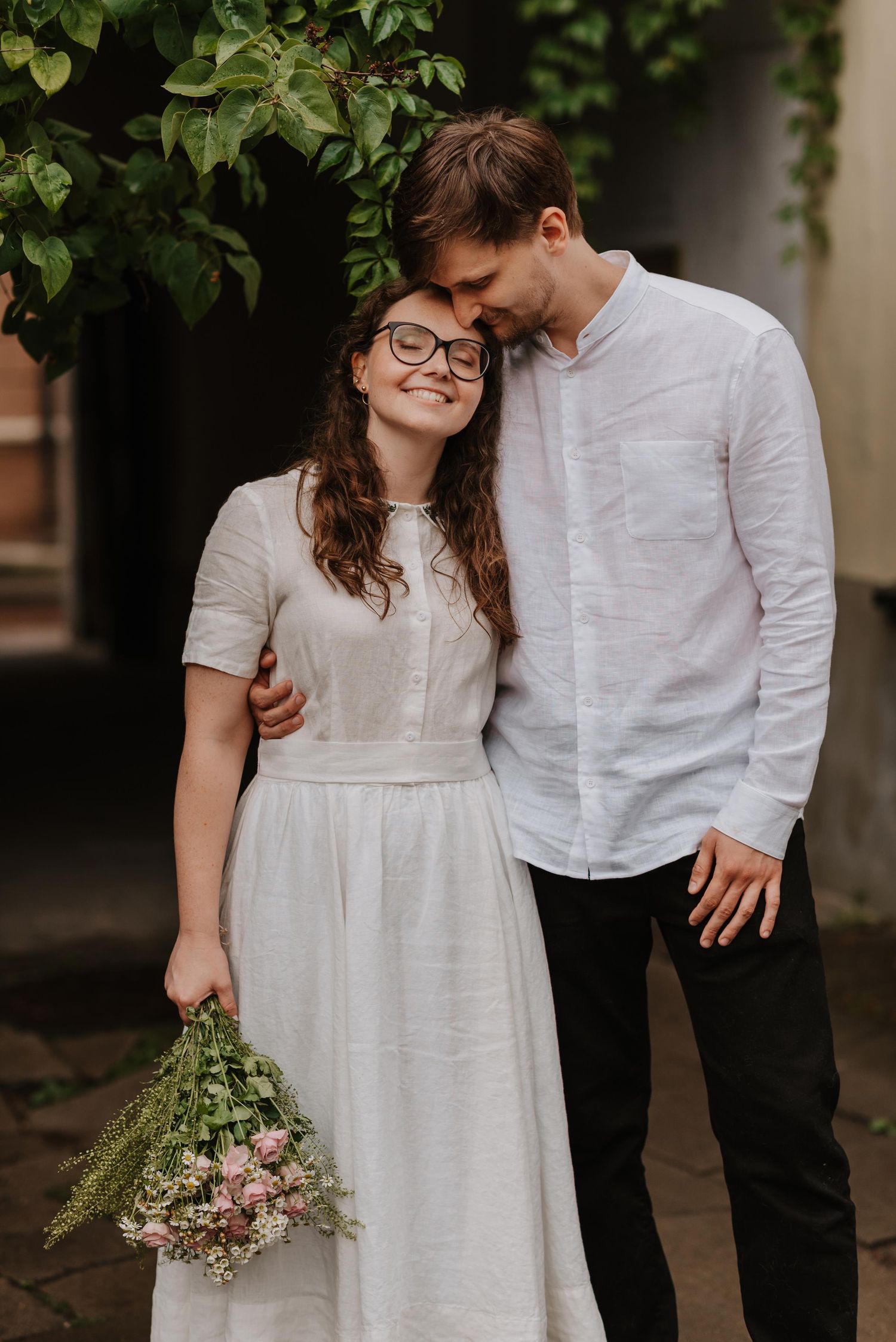 Romantic outdoor photoshoot features couple in white linen clothing surrounded by lush greenery.