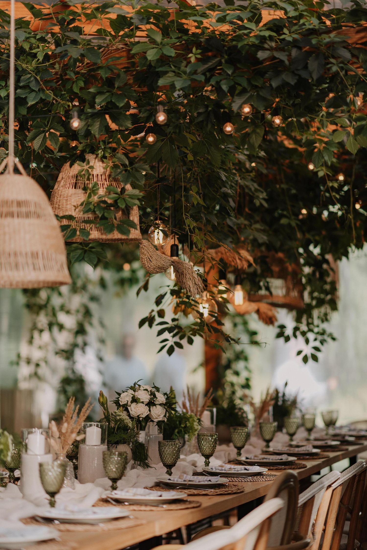 Wedding reception table decorated with greenery, hanging lights, and elegant place settings.