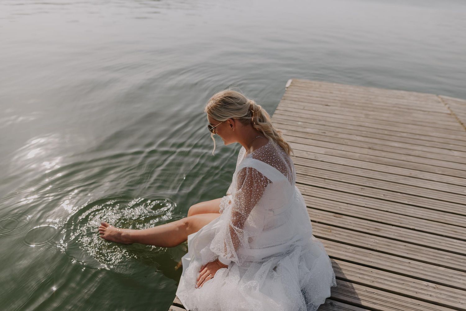 Person in white dress sits at edge of wooden dock by serene lake.