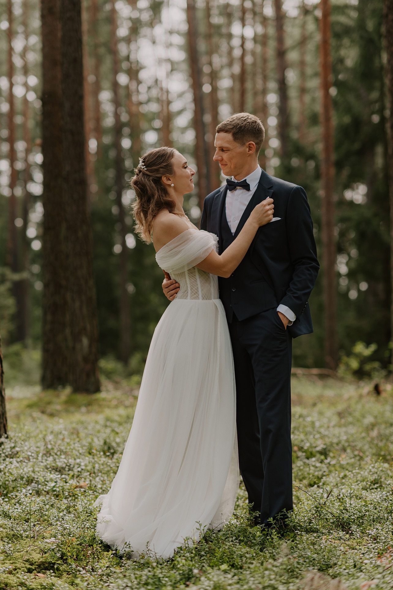 Elegant couple in wedding attire share an intimate moment in a pine forest.