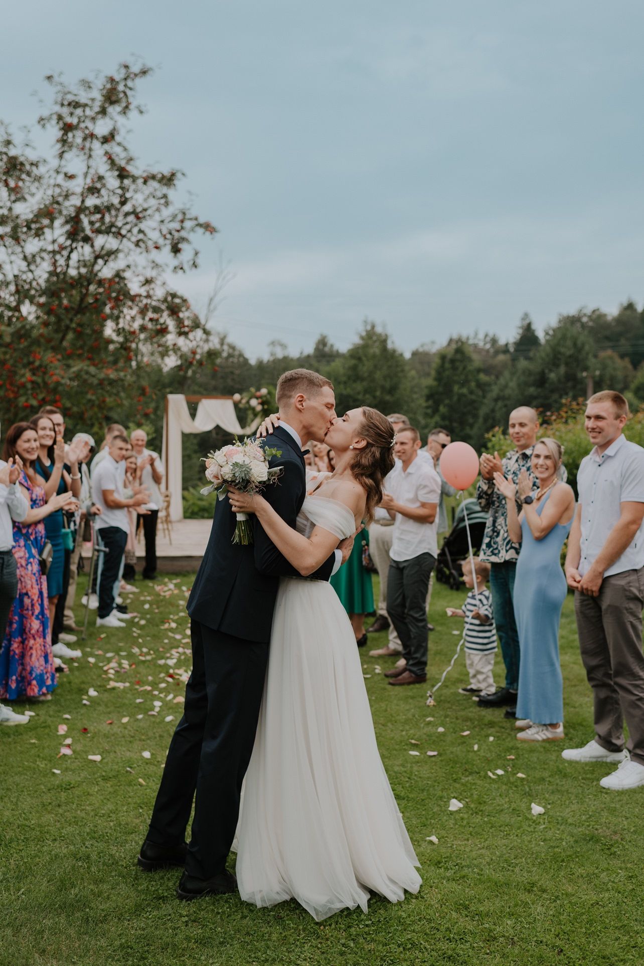 Newlyweds share their first dance surrounded by guests on a petal-covered lawn.