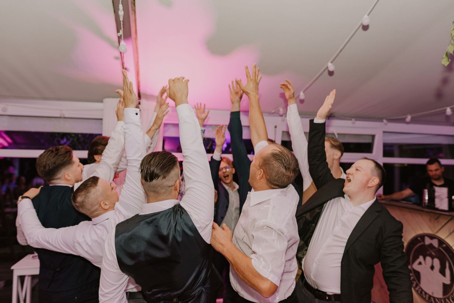 Energetic group dance celebration with raised arms at an indoor wedding reception.